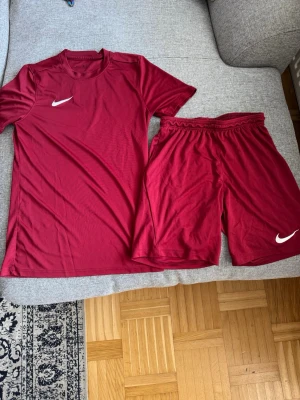Vinrött träningsset från Nike - Säljer ett vinrött träningsset från Nike med t-shirt och shorts. Båda plaggen har vit Nike-logga och är gjorda i ett lätt, snabbtorkande material som passar perfekt för träning eller sportaktiviteter.