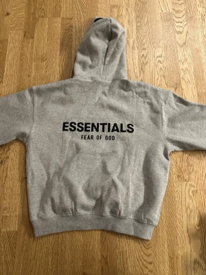 Grå Essentials Fear of God hoodie - Säljer en grå hoodie från Essentials Fear of God med svart logga på bröstet och stor text på ryggen. Klassisk känguruficka framtill och ribbade muddar. Tillverkad i mjukt bomullsmaterial och har en stor huva för extra komfort.