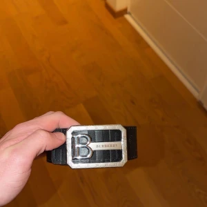 Svart Burberry bälte med metallspänne - Snyggt svart bälte från Burberry med ett stort, silverfärgat metallspänne där ett B och Burberry-loggan syns tydligt. Bältet har ett randigt mönster och känns riktigt lyxigt och stilrent. Perfekt accessoar för att lyfta din outfit. 