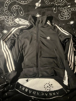 Svart Adidas track jacket med vita ränder - Klassisk svart track jacket från Adidas med vita tre ränder längs ärmarna och dragkedja framtill. Jackan har hög krage, två fickor med dragkedja och Adidas logga på bröstet. Perfekt för sport eller streetwear-stil. Lappen säger M men sitter mer som en S 🌞 