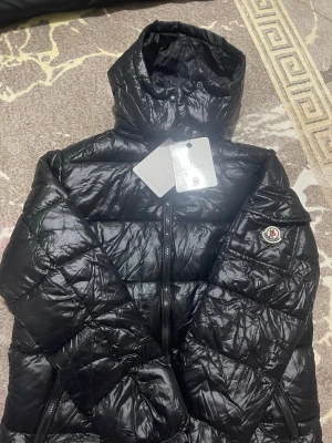Svart dunjacka från Moncler - Snygg svart dunjacka från Moncler med glansig finish och hög krage. Jackan har dragkedja framtill, två sidofickor och Moncler-logga på ärmen. Perfekt för kalla vinterdagar och riktigt trendig look.
