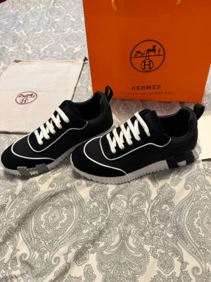 Svarta Hermes sneakers med vita detaljer - Snygga svarta sneakers från Hermès med vita kontrastsömmar och vita skosnören. Skorna har en unik sula med Hermès-logga undertill och coola detaljer i svart, vitt och grått. Tillverkade i en mix av mocka och textil för en modern look.