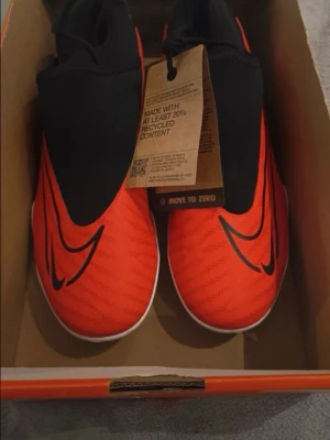 Nike röda fotbollsskor med dobbar - Snygga fotbollsskor från Nike i rött och svart med stora Swoosh-loggor. Skorna har en strömlinjeformad design, elastisk strumpa och vita sulor med svarta dobbar för bra grepp på planen. Tillverkade med minst 20% återvunnet material. Perfekta för dig som vill sticka ut på fotbollsplanen.