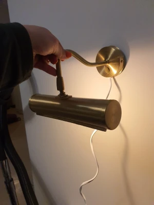 Mässingfärgad vägglampa - Snygg vägglampa i mässingfärgad metall med rund väggplatta och böjd arm. Lampan har en avlång, cylinderformad skärm och vit sladd för elanslutning. Perfekt som läslampa eller för att belysa tavlor.