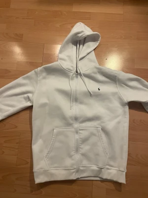 Vit hoodie från Polo Ralph Lauren - Säljer en clean vit hoodie från Polo Ralph Lauren med dragkedja framtill och klassisk huva med dragsko. Tröjan har två stora fickor fram och den ikoniska lilla mörkblå loggan broderad på bröstet. Perfekt till chill vardagsstil.