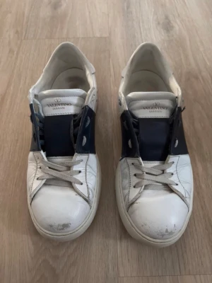 Valentino Open Sneakers - Snygga vita sneakers från Valentino Garavani med mörkblå detaljer på sidorna och tungan. Klassisk låg modell med vita snören och gummisula. Märkeslogga på plösen och under sulan. Perfekta för dig som vill ha en stilren och exklusiv look. Priset är såklart diskuterbart. Skorna passar till alla väder