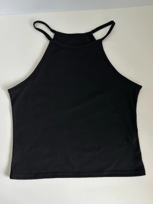 Svart halterneck-linne, storlek L - Fin svart halternecktopp / linne med hög halsringning och ärmlös design. Lika snygg till jeans som till kjol. Storlek L. Mycket gott skick, som ny.   Skickas mot porto, kan även hämtas i Stockholm. 