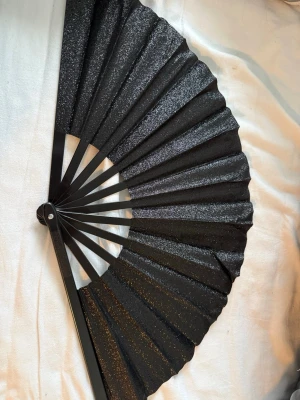 Svart solfjäder med glittereffekt - Snygg svart solfjäder med glittrande yta som ger en extra touch. Solfjädern är tillverkad av tyg och plast, och är lätt att fälla ihop och ta med sig. Perfekt accessoar för att sticka ut och hålla sig sval under varma dagar. Den är stor!