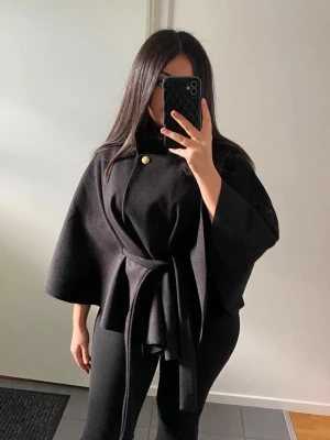 Svart cape-kappa med knytband - Stilren svart cape-kappa med vida ärmar och knytband i midjan. Kappan har en hög krage och en dekorativ guldig knapp framtill. Perfekt för dig som gillar en trendig och elegant look.