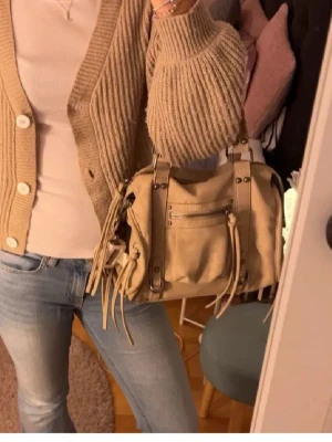 Beige axelremsväska med nitar - Balenciaga liknande väska från bershka som inte säljs längre💕💕Snygg beige handväska i mjukt material med dekorativa nitar och dragkedjor framtill. Väskan har långa fransar på dragkedjorna och justerbar axelrem. Perfekt för dig som gillar en avslappnad men trendig stil.