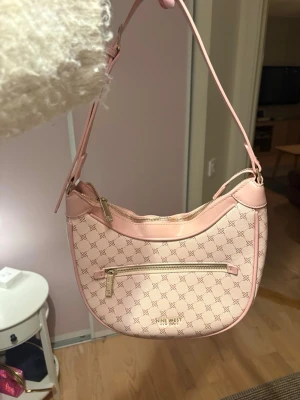Ljusrosa axelväska från Nine West - Snygg ljusrosa axelväska från Nine West med guldfärgade dragkedjor och logomönster över hela väskan. Väskan har en justerbar axelrem och en praktisk ytterficka med dragkedja. Tillverkad i konstläder med stilren, halvmåneformad design. Nypris: 600 kr! Aldrig använd
