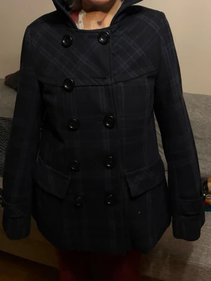 Mörkblå rutig rock med huva - Snygg mörkblå rock med rutigt mönster och dubbelknäppning framtill. Rocken har en huva, stora fickor och dekorativa knappar vid ärmsluten. Passar perfekt för dig som gillar en klassisk men ändå ungdomlig stil.