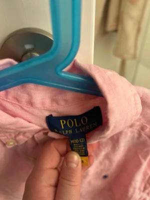 Rosa linneskjorta från Polo Ralph Lauren storlek 10-12 år - Säljer en klassisk rosa skjorta från Polo Ralph Lauren i linne. Skjortan har button-down krage, långa ärmar och den ikoniska broderade loggan på bröstet. Perfekt för en avslappnad men ändå stilsäker look.