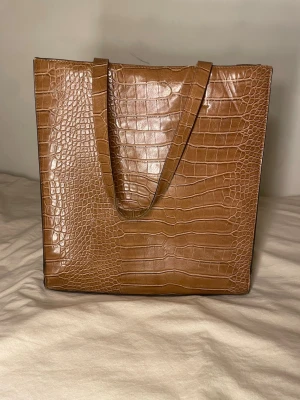 Brun shoppingväska i krokopräglat konstläder - Säljer en snygg shoppingväska i brunt krokopräglat konstläder. Väskan har en fyrkantig form och långa axelband som gör den enkel att bära. Perfekt för dig som vill ha en trendig och rymlig väska till vardagen.
