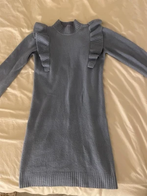 Ljusblå stickad klänning med volanger - Supersöt ljusblå stickad klänning med långa ärmar och volangdetaljer på axlarna. Ribbstickad halsringning och muddar vid ärmslut och nederkant. Perfekt för dig som vill ha en trendig och bekväm look i höst och vinter.