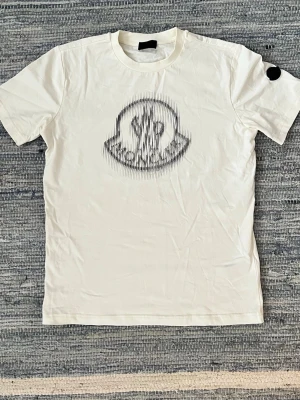 Vit Moncler t-shirt med logga - Säljer en vit t-shirt från Moncler med ett stort svart logotyptryck på bröstet i ett coolt, grafiskt mönster. Klassisk rund hals och korta ärmar. Perfekt för dig som gillar stilrena plagg med en twist. PRIS KAN DISKUTERAS