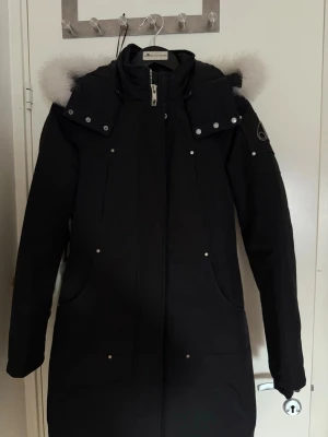 Svart dunjacka med pälskrage från Moose Knuckles - Stilren figursyd svart jacka med luva och vit pälskant som ger en exklusiv look. Silverfärgade detaljer och logotyp på vänster ärm som snygg accent. Endast använd ett fåtal gånger – i princip nyskick. Perfekt för kalla dagar när du vill vara både varm och välklädd. Nypris: 10 999kr Öppen för bud vid snabb affär.