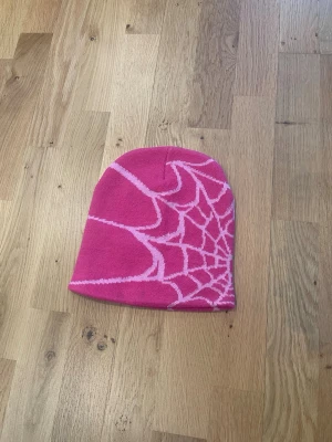 Rosa mössa spindelnät  - Rosa mössa med coolt ljusrosa spindelnätsmönster. Riktig statement-piece som passar perfekt till Y2K, alt- och streetwear-stil. Mjuk, stretchig och bekväm, sitter bra på huvudet.