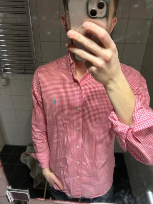 Rutig rosa skjorta från Ralph Lauren - Säljer en klassisk rosavitrutig skjorta från Ralph Lauren med button-down krage och blå logga broderad på bröstet. Skjortan har långa ärmar och knappar hela vägen framtill. Perfekt för en clean och stilren look.