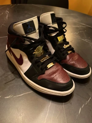 Air Jordan 1 Mid Beetroot - Snygga Air Jordan 1 Mid sneakers i färgerna bordeaux, svart och vitt. Skorna har klassisk Nike Swoosh, gulddetaljer på snörningen och perforerad tåbox. Tillverkade i läder med textildetaljer och ikonisk Air Jordan-logga på sidan.