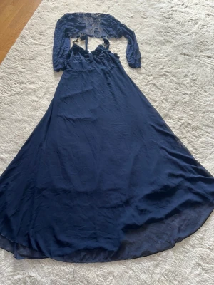 Marinblå långklänning med spets - Säljer en marinblå långklänning med spetsdetaljer upptill och långa ärmar. Klänningen har öppen rygg och v-ringad urringning, samt en flowy kjol i lätt material. Perfekt för dig som vill ha en elegant look med unika detaljer.