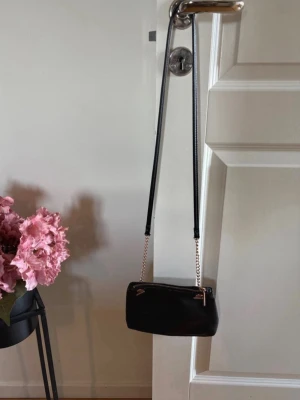 Svart axelväska med guldkedja - Snygg svart axelväska i slätt konstläder med guldfärgade kedjedetaljer och dragkedja upptill. Väskan har en stilren, rektangulär form och passar perfekt för dig som vill ha en enkel men trendig accessoar till din outfit. (Finns ett litet märke se bild) 