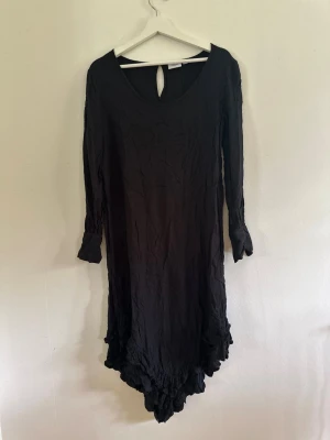 Svart långklänning från Saint Tropez XS - Svart långklänning från Saint Tropez med lång ärm och asymmetrisk volangkant nedtill. Klänningen har en enkel, stilren design med rund halsringning och knappdetalj i nacken. Perfekt för dig som gillar en bohemisk och avslappnad look. Passar mig som är M