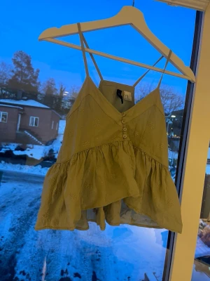 Beige broderad linnetopp med volang - Söt beige linnetopp med tunna axelband och broderade blommor. Toppen har v-ringning, knappar framtill och volangdetaljer som ger en luftig känsla. Perfekt för varma dagar och enkel att styla med jeans eller kjol.