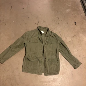 Olivgrön parkajacka med fickor - Snygg olivgrön parkajacka med fyra stora fickor framtill och klassisk krage. Jackan har långa ärmar och knäppning framtill. Perfekt för dig som gillar en avslappnad och stilren look. Storlek 158, passar som xxs-xs. Pris kan diskuteras vid snabb affär