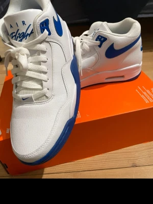 Nike Air Flight vita och blå sneakers - Säljer ett par Nike Air Flight sneakers i vitt med blå detaljer och swoosh. Skorna har en klassisk siluett med lågt skaft, vita skosnören och blå sula. Ovandelen är i läder och mesh för en sportig look. Perfekta för dig som gillar retrostil och streetwear.