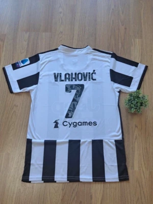 Juventus matchtröja Vlahović #7 - Juventus matchtröja i svartvitt med Vlahović och nummer 7 på ryggen. Klassiska breda ränder, kort ärm och tryckta loggor från Cygames och Serie A på ärmen. Tillverkad i lätt och ventilerande material, perfekt för fotbollsträning eller match.