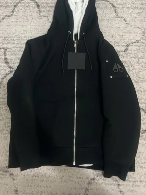 Svart hoodie från Moose Knuckles - Säljer en svart hoodie från Moose Knuckles med dragkedja framtill och vit fluffig insida. Hoodien har en stor huva med dragsko och logotyp på ärmen i metall. Perfekt för dig som vill ha en stilren och exklusiv look.