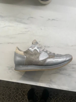 Philippe Model silvriga sneakers - Snygga sneakers från Philippe Model i silver och beige med metallicdetaljer och tygpaneler. Klassisk låg modell med gummisula och logotyp på sidan. Perfekta för dig som vill sticka ut med stilrena och trendiga skor.