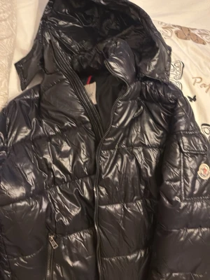 Svart dunjacka från Moncler med huva - Snygg svart dunjacka från Moncler med glansig finish och fast huva. Jackan har quiltad design, dragkedja framtill och en Moncler-logga på ärmen. Perfekt för kalla vinterdagar och riktigt trendig look.