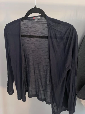 Mörkblå tunn kofta Street One - Tunn och luftig mörkblå kofta från Street One. Koftan har öppen front utan knappar, långa ärmar och är tillverkad i ett lätt, transparent material som ger en avslappnad look. Perfekt att slänga över en topp för en enkel och stilren outfit.