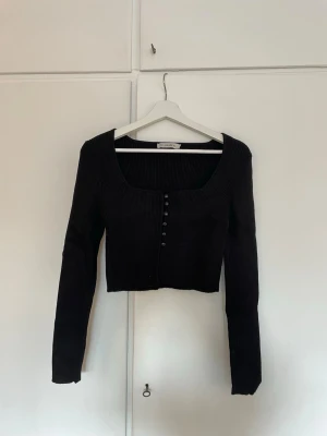 Svart ribbad tröja med knappar - Svart ribbad kofta/tröja med lång ärm och croppad passform. Djup fyrkantig ringning och knäppning framtill med små svarta knappar. Perfekt att styla till jeans eller kjol för en cool look.