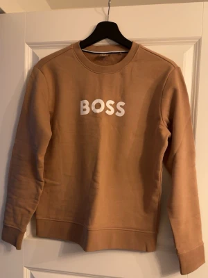 Beige sweatshirt från BOSS - Säljer en beige sweatshirt från BOSS med vit logotyp på bröstet. Tröjan har rund halsringning, långa ärmar och ribbade muddar vid ärmslut och nederkant. Perfekt för en avslappnad och stilren look.