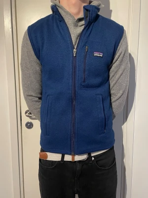 Patagonia fleeceväst - Snygg blå fleeceväst från Patagonia med dragkedja framtill och hög krage. Västen har två sidofickor och en bröstficka med dragkedja. Perfekt att slänga över en hoodie för extra värme. Klassisk logga på bröstet.