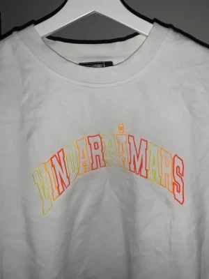 Hov1 tröja vindar på mars  - Vit t-shirt med texten 'VINDARPÅMARS' i stora bokstäver framtill, tryckt i färgerna rött, gult och orange. Rund halsringning och klassisk passform. 