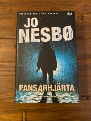 Pansarhjärta av Jo Nesbø - Spännande thrillerroman av Jo Nesbø om polisen Harry Hole. Boken har hårda pärmar med ett omslag i blått och svart, där en siluett står i regnet. Texten är vit och boken är översatt av Per Olaisen. Perfekt för dig som gillar kriminalromaner och nordisk noir.