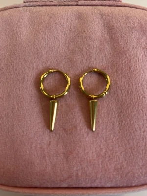 Guldpläterade hoops med spike - Säljer ett par coola örhängen i guldpläterad metall. De har en rund hoop-design med en spetsig spike som hänger ned. Perfekt för dig som gillar edgy och trendiga accessoarer. Passar till många olika stilar och är lätta att bära.