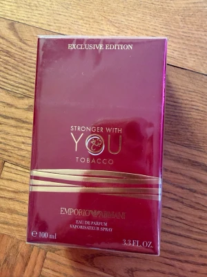 Stronger With You Tobacco parfym - Stronger With You Tobacco från Emporio Armani är en exklusiv parfym i en elegant mörkröd kartong med guldfärgade detaljer. Förpackningen rymmer 100 ml Eau de Parfum och har en lyxig och modern design som verkligen sticker ut.