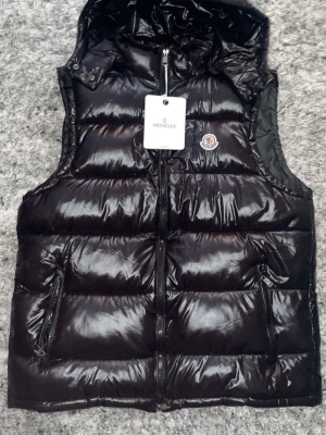 Svart dunväst från Moncler - Snygg svart dunväst från Moncler med glansig finish och klassisk logga på bröstet. Västen har dragkedja framtill, två fickor med dragkedja och huva. Perfekt för lager-på-lager och streetstyle.