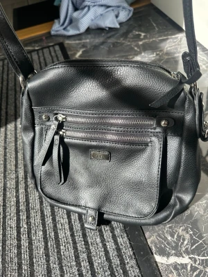 Svart axelväska från Feel - Snygg svart axelväska från Feel i mjukt konstläder. Väskan har två dragkedjefickor framtill, justerbar axelrem och silvriga metalldetaljer. Perfekt för dig som vill ha en stilren och praktisk väska med plats för det viktigaste.
