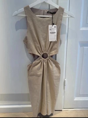 Beige omlottklänning från ZARA - Trendig beige klänning från ZARA med snygg cut-out och rynkad detalj i midjan. Ärmlös modell med rund halsringning och omlottkänsla. Perfekt för dig som gillar stilrena och moderna plagg.