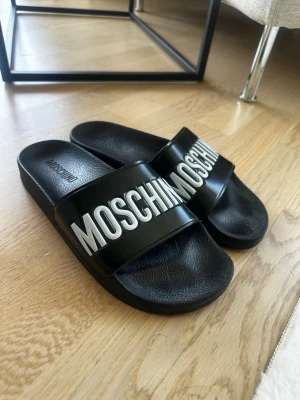 Svarta Moschino tofflor strl 41 - Snygga svarta badtofflor från Moschino med bred rem och stor vit logotyp framtill. Tillverkade i syntetmaterial med en klassisk slip-on design och bekväm fotbädd. Perfekta för en trendig och avslappnad stil. Köpta på Miinto. Nypris ca: 1700kr 