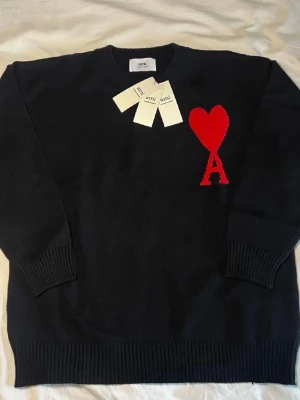 Svart stickad tröja från Ami - Svart stickad tröja från Ami Alexandre Mattiussi med röd hjärtlogga och bokstaven A på bröstet. Tröjan har rund halsringning, ribbade muddar och är gjord i mjukt material. Perfekt för dig som gillar stilrena plagg med en twist.