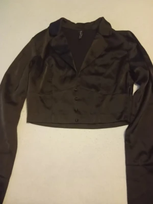 Svart satin blus med knappar - Snygg svart blus i glansigt satinmaterial med figurnära passform. Blusen har långa ärmar, klassisk krage och fyra klädda knappar framtill. Perfekt för dig som vill ha en stilren och trendig look.