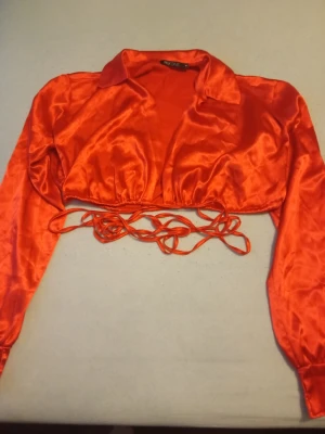 Röd croppad omlottopp i satin - Snygg röd croppad omlottopp från NEYONE i glansigt satinmaterial. Toppen har långa ärmar, krage och knytband i midjan för en trendig look. Perfekt för dig som vill sticka ut med en färgstark och modern stil.
