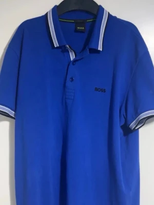 BOSS Piké Blå - ‼️Med short 900kr.                                                              Snygg blå pikétröja från BOSS med vit och mörkblå rand på krage och ärmslut. Tröjan har klassisk krage, knappar framtill och broderad BOSS-logga på bröstet. Regular fit för en bekväm passform.